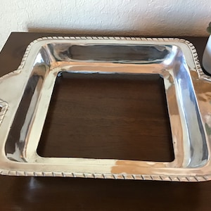 May include: A silver serving tray with a square shape and a raised rim. The tray has a decorative rope-like design around the edge.