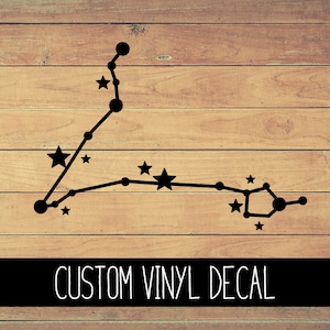 May include: Black vinyl decal of the Pisces constellation on a wood background. The decal features a stylized outline of the constellation with small stars. The text "CUSTOM VINYL DECAL" is printed below the constellation.