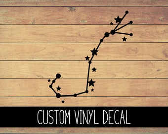 May include: Black vinyl decal of the constellation Scorpio with stars and lines on a wood background. The text "CUSTOM VINYL DECAL" is below the constellation.