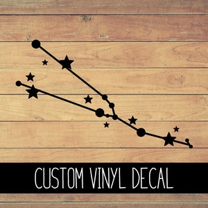 May include: Black vinyl decal of the Taurus constellation, featuring stars and connected dots, against a wooden background. The text "CUSTOM VINYL DECAL" is displayed in white on a black banner.