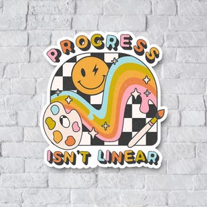 May include: A white sticker with a rainbow, a smiley face, a paint palette, and a paintbrush. The text on the sticker reads "Progress isn't linear".