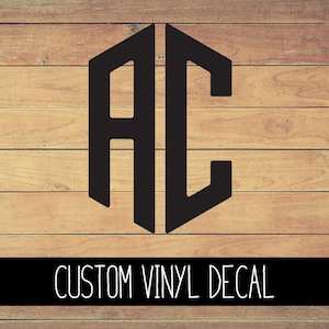 May include: A black vinyl decal with the letters "AC" in a geometric shape. The decal is on a wood background. CUSTOM VINYL DECAL is written in white text below the decal.