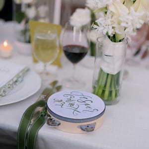 May include: A white tambourine with a green ribbon and the text "And See's God's Gift" written in black calligraphy. The tambourine is on a white tablecloth with a white flower bouquet in a glass vase.