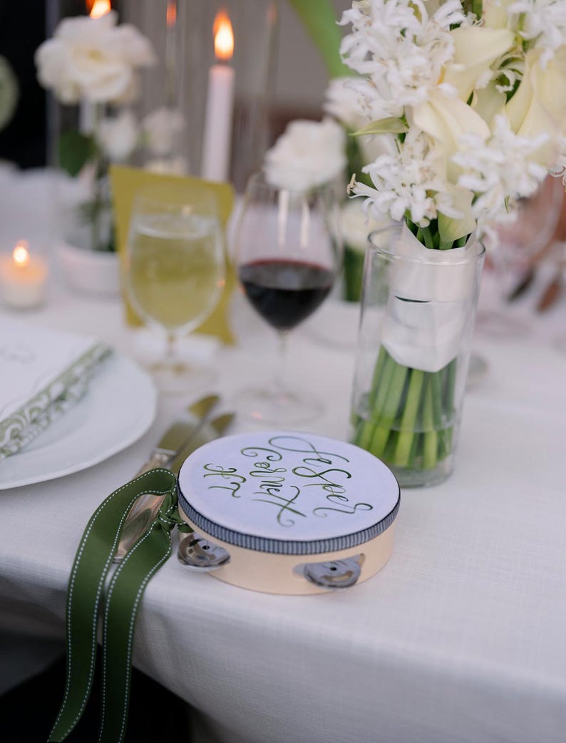 May include: A white tambourine with a green ribbon and the text "And See's God's Gift" written in black calligraphy. The tambourine is on a white tablecloth with a white flower bouquet in a glass vase.