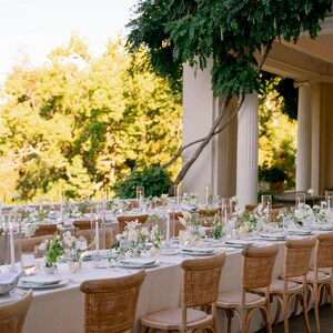May include: A long, white tablecloth with a row of light brown wicker chairs. The table is set with white plates, clear glasses, and white flowers in glass vases. The table is set up outdoors on a patio with a view of green trees.