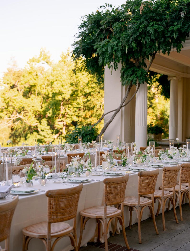 May include: A long, white tablecloth with a row of light brown wicker chairs. The table is set with white plates, clear glasses, and white flowers in glass vases. The table is set up outdoors on a patio with a view of green trees.