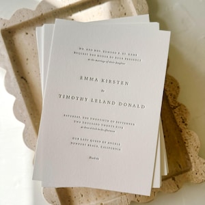 May include: A stack of white wedding invitations with black text, resting on a beige, scalloped-edge tray. The invitation text includes names, date, time, and location: Our Lady Queen of Angels, Newport Beach, California.