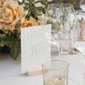 May include: A white card with the text "table 16" written in calligraphy, sitting on a wooden stand. The card is on a white tablecloth with a glass of water and a wine glass in the background.