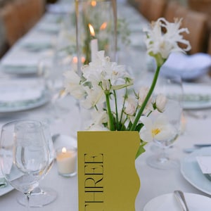 May include: A white tablecloth with a yellow table number card that says "THREE" on it. White flowers in a vase are in the center of the table. There is a clear glass candle holder with a white candle in it.