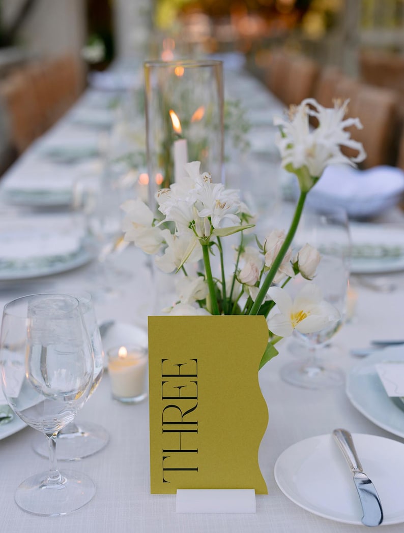 May include: A white tablecloth with a yellow table number card that says "THREE" on it. White flowers in a vase are in the center of the table. There is a clear glass candle holder with a white candle in it.