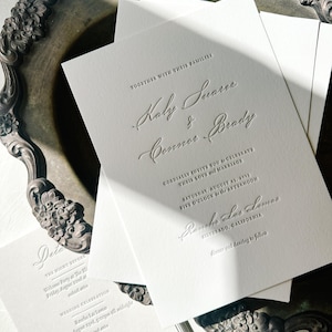 May include: Elegant white wedding invitations with embossed text, including names "Kaly Suarez & Connor Brady" and event details. The invitations are arranged on a decorative silver tray, showcasing a classic and sophisticated design.
