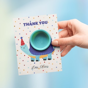 May include: A thank you card featuring a cartoon turtle with a party hat and a turquoise container. The card has the text "THANK YOU" and "Love, Oliver" with a polka dot design. The container is a circular shape.