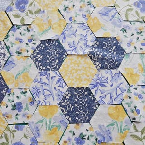 May include: Close-up of a patchwork of hexagonal fabric pieces. The fabrics feature floral patterns in yellow, blue, and white. Some pieces have solid yellow or navy blue designs. The arrangement suggests a quilt or craft project.