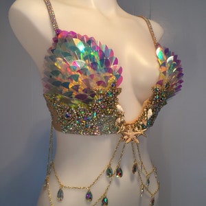 May include: A gold and iridescent mermaid-themed bra with a seashell design, rhinestones, and a starfish accent. The bra has a chain with teardrop-shaped crystals hanging from it.