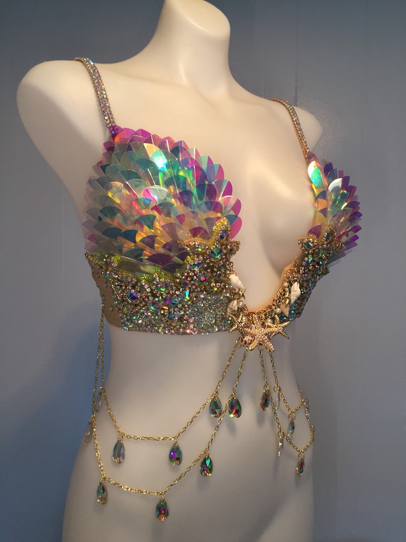 May include: A gold and iridescent mermaid-themed bra with a seashell design, rhinestones, and a starfish accent. The bra has a chain with teardrop-shaped crystals hanging from it.