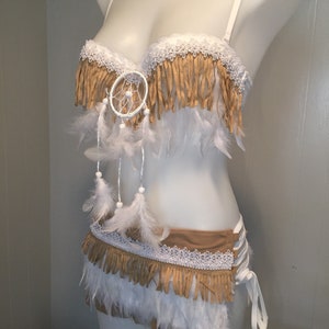 May include: A white bra and skirt set with tan fringe, white lace trim, and white feathers. A dreamcatcher is centered on the bra. The skirt has a lace-up side detail. The set is displayed on a mannequin.