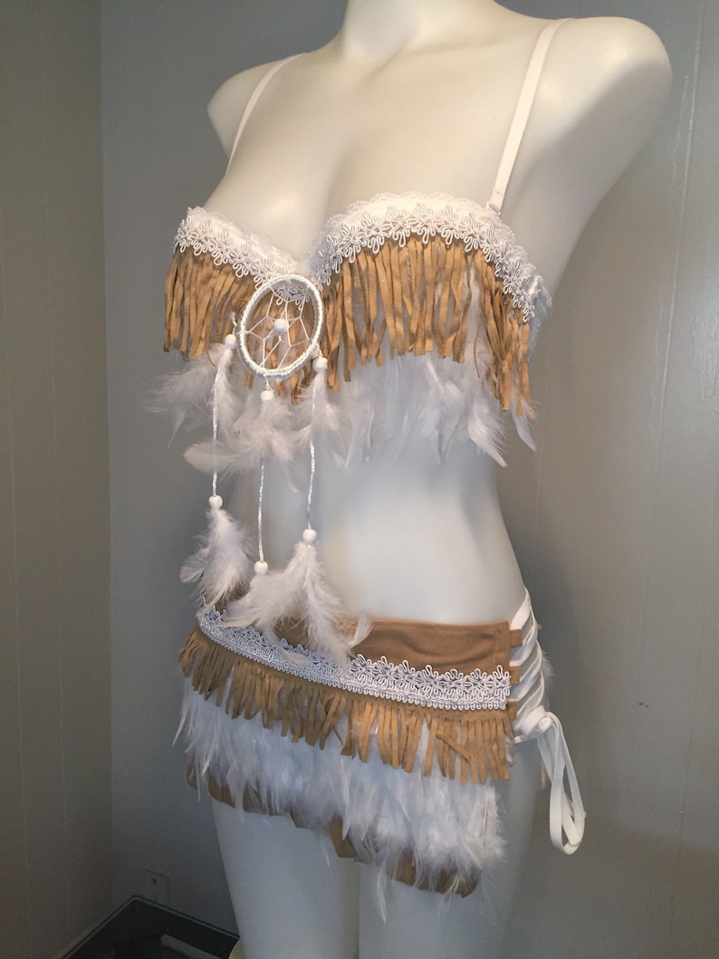 May include: A white bra and skirt set with tan fringe, white lace trim, and white feathers. A dreamcatcher is centered on the bra. The skirt has a lace-up side detail. The set is displayed on a mannequin.