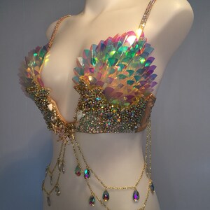 May include: A gold and iridescent bra with a mermaid-inspired design. The bra features a shell-shaped cup with iridescent scales and a chain with teardrop-shaped crystals.