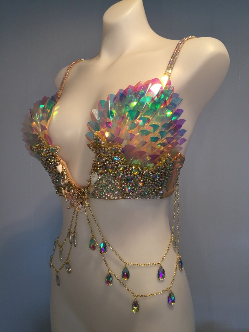 May include: A gold and iridescent bra with a mermaid-inspired design. The bra features a shell-shaped cup with iridescent scales and a chain with teardrop-shaped crystals.