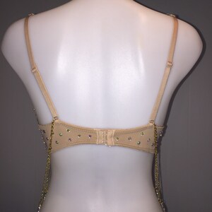 May include: A nude-colored bra with adjustable straps and a back closure. The bra is decorated with iridescent rhinestones and has gold chains hanging from the bottom.
