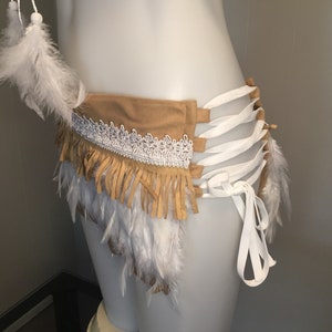 May include: A tan suede belt with white lace trim and fringe, adorned with white feathers. The belt features white ribbon ties and is designed to be worn around the waist. The overall style is bohemian.