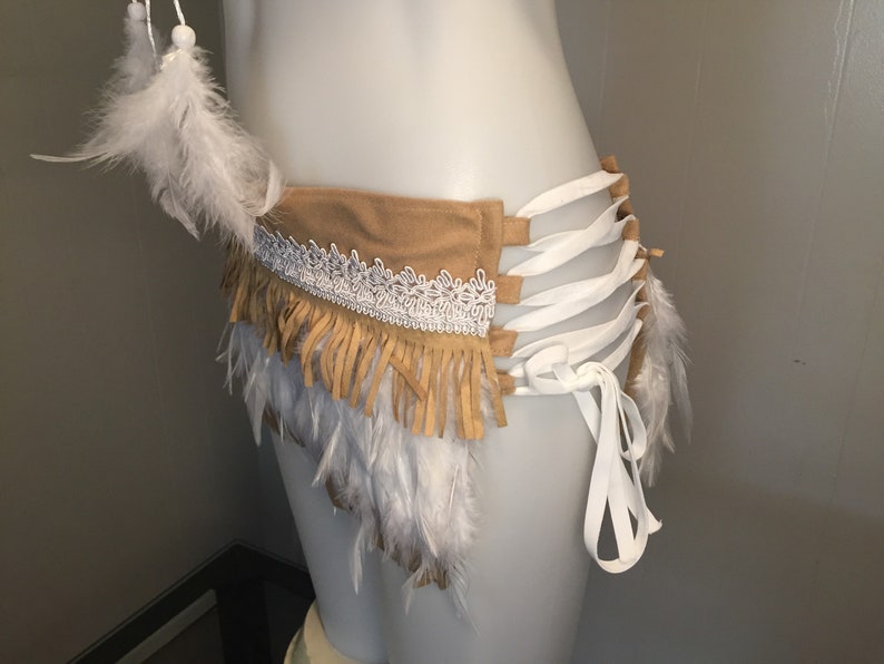 May include: A tan suede belt with white lace trim and fringe, adorned with white feathers. The belt features white ribbon ties and is designed to be worn around the waist. The overall style is bohemian.