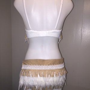 May include: A white lace bra and a matching skirt with a tan suede waistband, white lace trim, and tan fringe. The skirt has a layer of white feathers at the bottom. The outfit is displayed on a white mannequin.