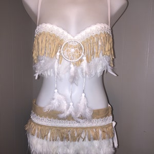 May include: A two-piece costume set on a mannequin. The top is a bra with fringe and a dreamcatcher. The bottom is a skirt with fringe and feathers. The costume is tan, white, and cream-colored.