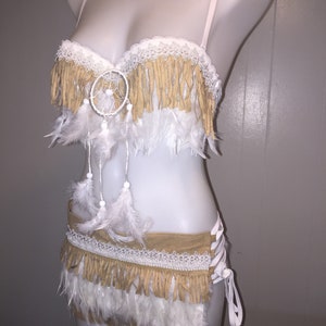 May include: A two-piece cream-colored lingerie set with a Native American theme. The top and bottom feature tan fringe, white lace, and white feathers. A dreamcatcher is centered on the top. The set is displayed on a mannequin.