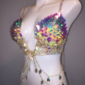 May include: A iridescent, multi-colored, sequined bra with gold chains and teardrop-shaped crystals. The bra has a mermaid-inspired design.