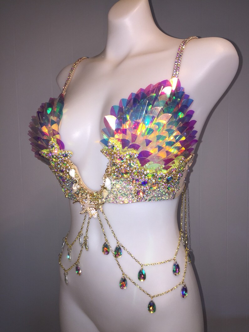 May include: A iridescent, multi-colored, sequined bra with gold chains and teardrop-shaped crystals. The bra has a mermaid-inspired design.