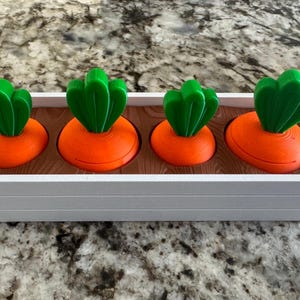 May include: A white rectangular planter box with four orange carrot-shaped decorations. Each carrot has green leafy tops. The carrots sit in a brown wooden base within the white box. The planter is on a speckled countertop.
