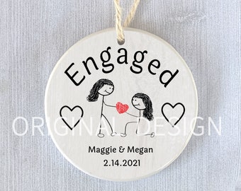 May include: White ceramic ornament with the word "Engaged" in black lettering. Two stick figures are holding a red heart. The text "Maggie & Megan 2.14.2021" is below the figures.