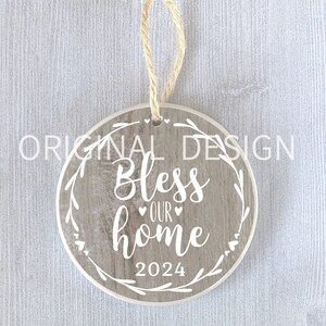 Op de afbeelding: Ronde houten ornament met een rustiek houtnerf ontwerp. Het heeft een witte krans met de tekst "Bless our home 2024" in een wit cursief lettertype.