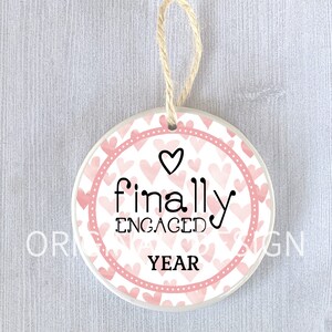 Könnte beinhalten: Ein weißer Keramik-Anhänger mit einem rosafarbenen Aquarell-Herz-Muster. Der Anhänger hat einen rosafarbenen Rand mit weißen Punkten und den Text "finally engaged YEAR" in Schwarz.