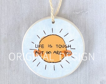 May include: A round ceramic ornament with a blue background and a yellow sun with the text "Life is tough but so are you".
