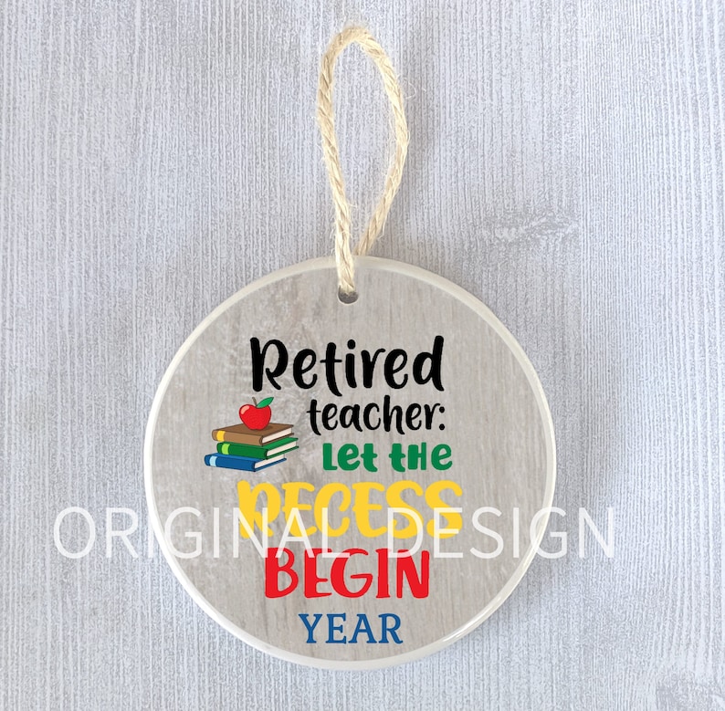 Op de afbeelding: Een ronde, witte keramische ornament met een houtnerfpatroon. Het heeft de tekst "Gepensioneerde leraar: Laat de pauze beginnen jaar" in zwarte letters met een rode appel en een stapel boeken. De ornament heeft een bruine touwhalter.