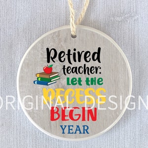 Op de afbeelding: Een ronde keramische ornament met een houtnerf achtergrond. De ornament heeft de tekst "Gepensioneerde leraar: Laat de pauze beginnen jaar" in zwart, groen, geel, rood en blauw. Een rode appel en een stapel boeken zijn afgebeeld boven de tekst.