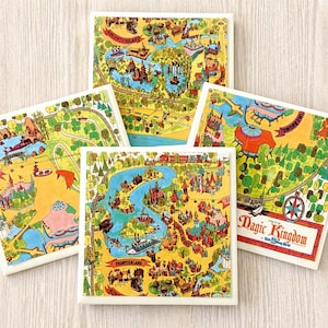 May include: Five square ceramic coasters with colorful vintage maps of a theme park. The maps feature illustrations of rides, attractions, and landmarks, with text that reads "Magic Kingdom" and "Frontierland". The coasters are set against a light wood background.