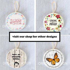 Op de afbeelding: Vier ronde keramische ornamenten met verschillende ontwerpen. De eerste ornament is wit met een roze bloemenkrans en de tekst "It's a Wonderful Life". De tweede ornament is wit met een roze, gele en blauwe bloemenkrans en de tekst "Strong as a Mother". De derde ornament is wit met een houtnerf achtergrond en de tekst "have an adequate holiday". De vierde ornament is wit met een zwart-witte bloemenkrans en een monarchvlinder.