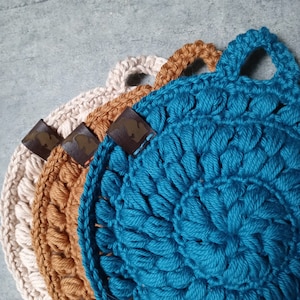 May include: Three crocheted pot holders in beige, brown, and teal with leather labels. The pot holders have a textured, bumpy surface.