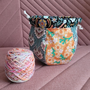 Peut inclure: Un bol à laine en tissu coloré avec une fermeture à cordon. Le bol est fait de tissu patchwork avec des motifs floraux et paisley dans des tons de turquoise, orange et blanc. Une pelote de laine multicolore se trouve à côté du bol.