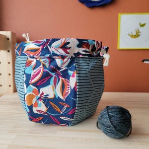 Puede incluir: Una bolsa de cordón azul y blanco con flores con un panel a rayas gris y blanco. La bolsa está llena de una bola de hilo azul.