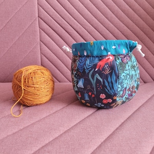 May include: A teal and black drawstring bag with a floral and animal print. The bag is filled with a ball of orange yarn.