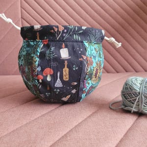 Puede incluir: Una bolsa de cordón negro con estampado floral y de pociones. La bolsa está llena de hilo y está sobre una superficie rosa texturizada.