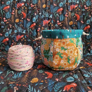 Puede incluir: Una bolsa de proyecto de tela colorida con una parte superior turquesa y un diseño de patchwork de patrones florales naranja, amarillo y verde. La bolsa está sobre un fondo oscuro con un patrón floral. También hay una bola de hilo rosa y blanco en la imagen.