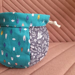 Puede incluir: Una bolsa de cordón de color turquesa y gris con un patrón de pájaros y flores blancas. La bolsa está hecha de tela y tiene un cordón de ajuste blanco.