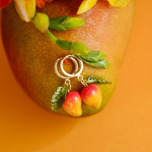 May include: Gold hoop earrings with mango charms. The earrings are hanging from a mango fruit.