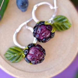 Puede incluir: Pendientes de aro plateados con abalorios de mora de cristal. Los abalorios presentan moras de color púrpura intenso con detalles negros y hojas verdes. Los pendientes descansan sobre una superficie marrón claro.