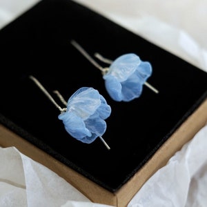 Puede incluir: Un par de delicados pendientes de flores azules con postes de plata, presentados en una caja negra forrada de terciopelo negro.